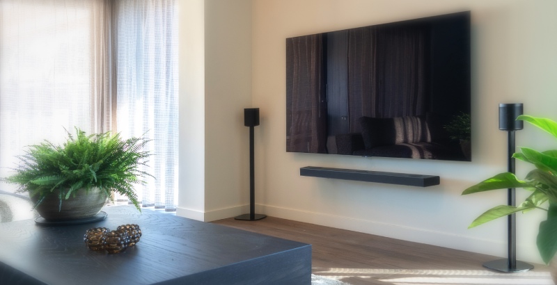 Comment installer des enceintes ? Découvrez tout ce qu'il faut savoir à ce sujet. Une télévision accrochée au mur avec deux enceintes à côté, posées sur des supports dans le salon.