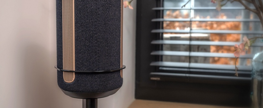 Gros plan d'une enceinte noire sur un support Vebos avec un support, placée dans un intérieur moderne près d'une fenêtre avec des stores.